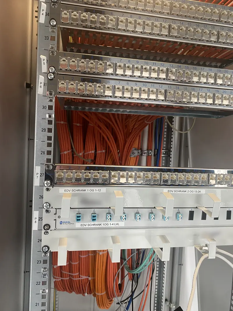 Netzwerkschrank mit beschrifteten Patchpanels, sauber gebündelter Datenverkabelung und Glasfaser-Verteilerfeld zwischen den Etagen