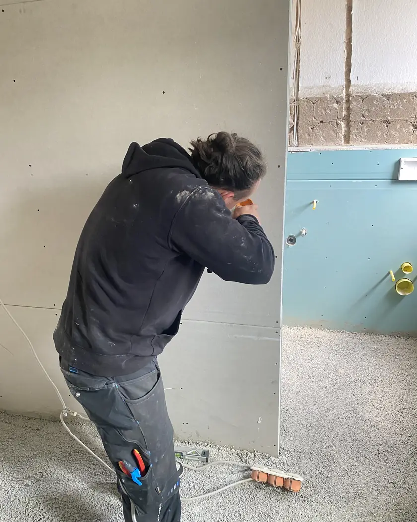 Monteur von ElektroTechnik Posengis setzt beim Umbau eine Hohlwanddose in eine neue Trockenbauwand, daneben eine Wand mit Feuchtraumplatten und vorbereiteten Leerrohren