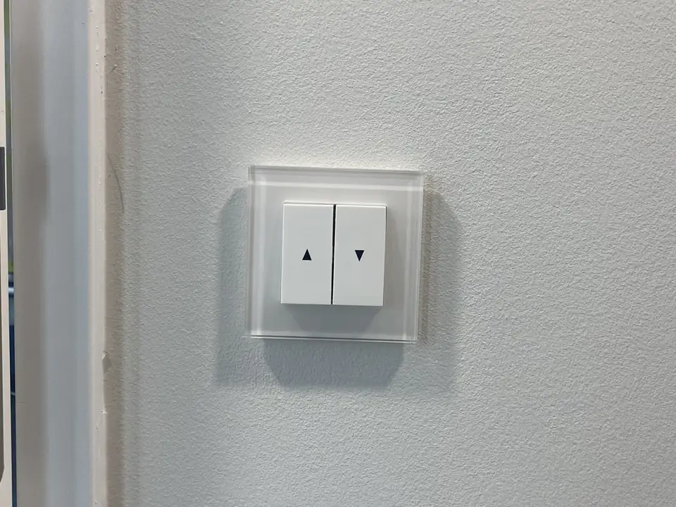 Jalousietaster mit Pfeiltasten in einem Glasrahmen an einer weißen Wand neben einem Türrahmen