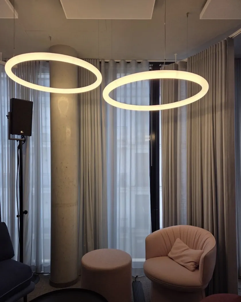 Zwei große LED-Ringpendelleuchten mit ruhigem, gleichmäßigem warmweißem Licht in einer Lounge mit Vorhängen und Sesseln