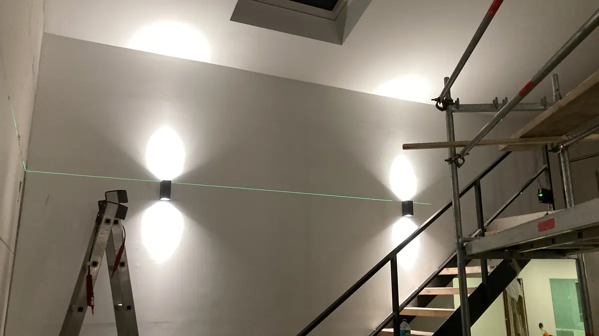 Montage von LED-Wandleuchten im Treppenhaus, ausgerichtet mit einem grünen Kreuzlinienlaser, daneben Leiter und Gerüst