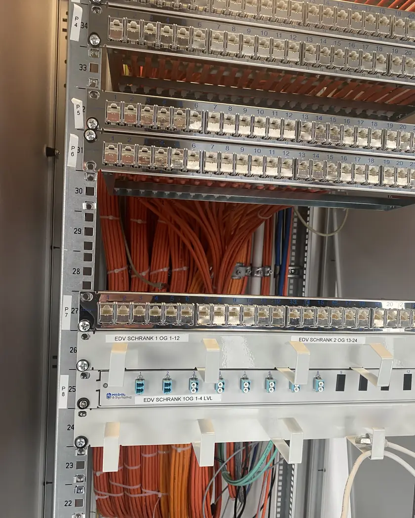 Offener Netzwerkschrank mit mehreren nummerierten Patchfeldern, dahinter gebündelte orangefarbene Datenkabel und darunter ein LWL-Patchfeld mit Glasfaserkupplungen