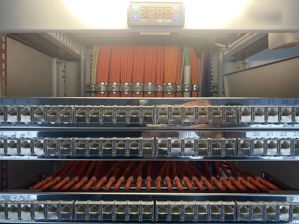 Drei Patchfelder mit nummerierten Netzwerkports, dahinter sauber gebündelte und befestigte orangefarbene Datenkabel, oben eine Temperaturanzeige der Lüftersteuerung