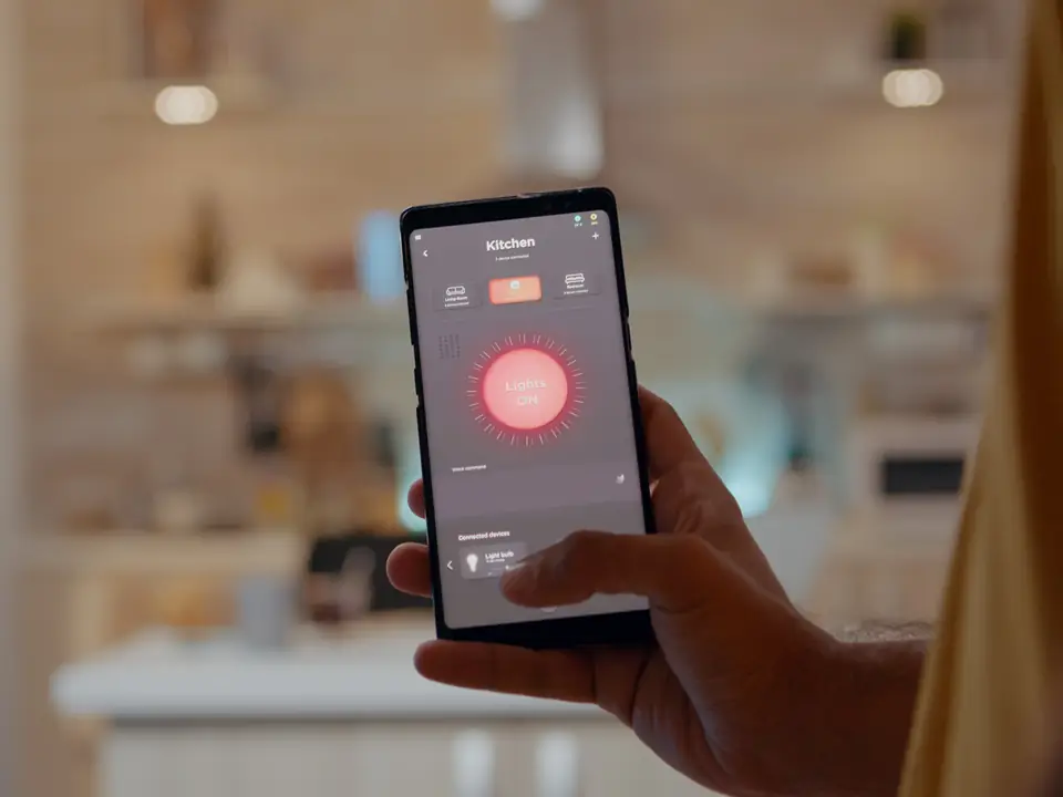 Hand hält ein Smartphone mit einer Smart-Home-App zur Lichtsteuerung, im Hintergrund eine unscharfe Küche