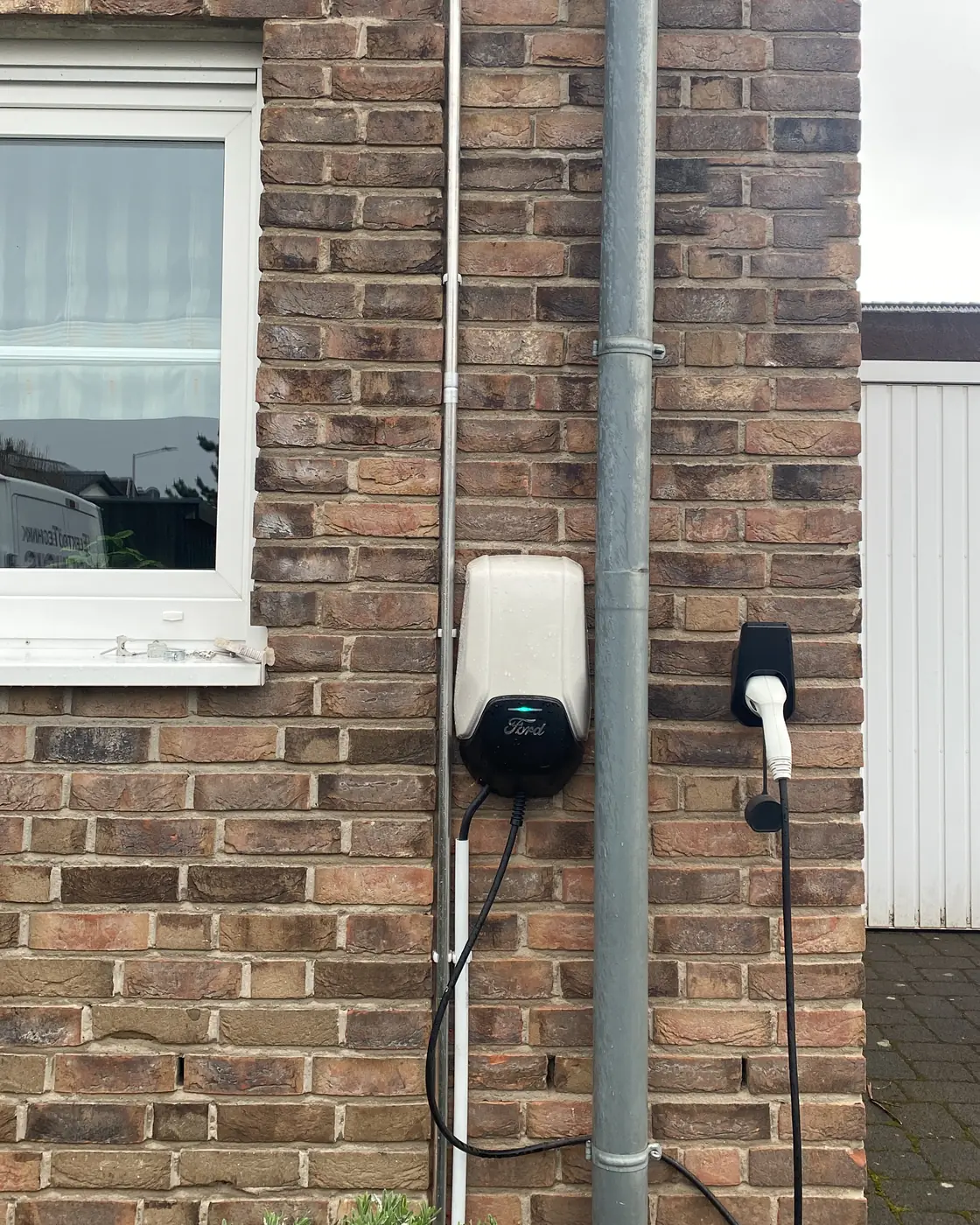 Wallbox an der Klinker-Außenwand eines Einfamilienhauses, Zuleitung sauber im Installationsrohr an der Fassade verlegt, daneben ein Halter für den Ladestecker