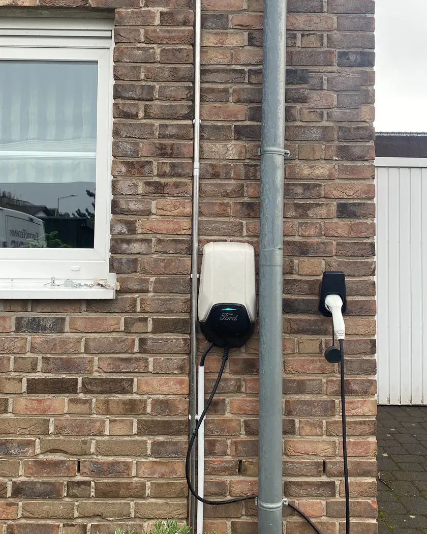 Wallbox an der Klinker-Außenwand eines Einfamilienhauses, Zuleitung sauber im Installationsrohr an der Fassade verlegt, daneben ein Halter für den Ladestecker