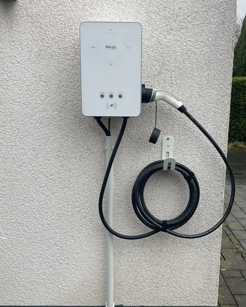 Wallbox mit fest angeschlossenem Typ-2-Ladekabel an einer verputzten Hauswand, Kabel aufgerollt auf einem Wandhalter, Zuleitung im weißen Rohr